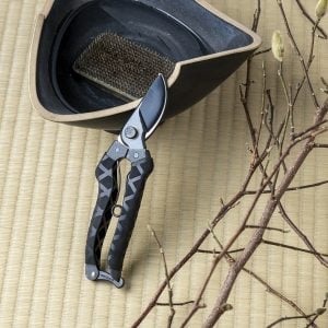 Black Masamune pruning shears resting against Japanese ceramic on tatami mat with styled branches nearby